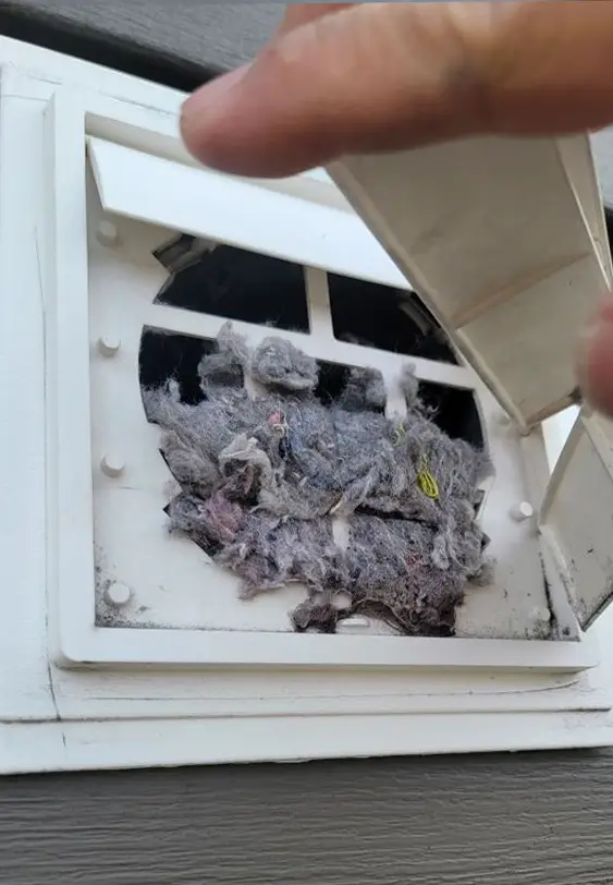 A dryer vent on the side of a building is clogged with a large accumulation of lint and debris. The best duct cleaning solution reveals itself as a hand lifts the partially open vent cover, exposing the blockage. The siding of the building is visible in the background, waiting for expert attention.