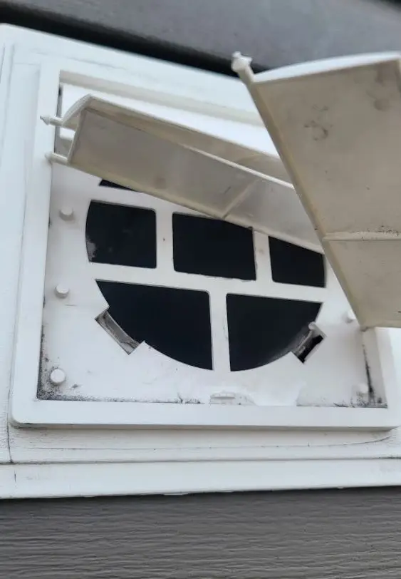 Close-up of a white vent on a gray wall. The vent has a square housing with an open flap revealing a circular interior with a grid-like structure. Dirt smudges are visible around the edges, highlighting the need for the best dryer vent cleaning near me to maintain efficiency and cleanliness.