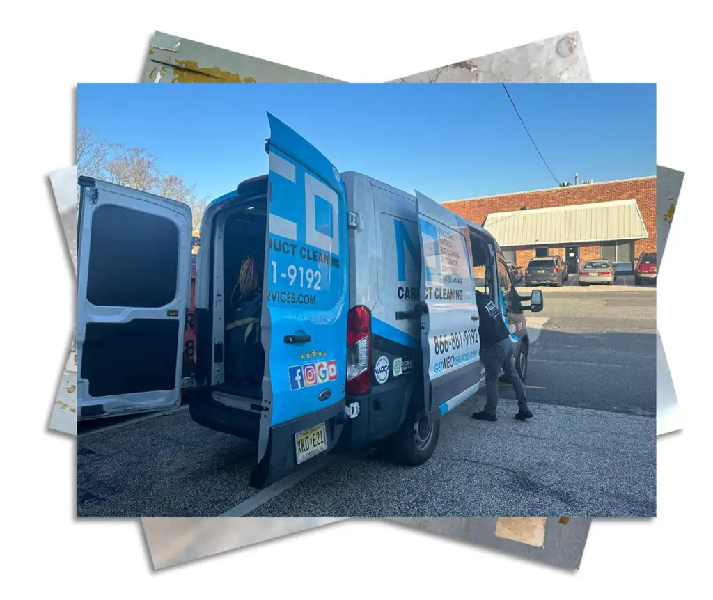 A blue and white van with DUCT CLEANING boldly displayed on the side is parked in a lot. The back doors are open, revealing one of New Jerseys best air duct cleaners moving equipment. The van showcases contact details, social media logos, and a license plate. A brick building stands in the background.