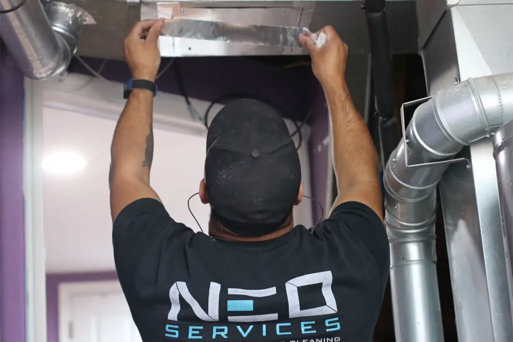 A person wearing a black cap and a NEO SERVICES T-shirt is carefully applying duct tape to a metallic duct in a building with purple walls. As one of the finest air duct cleaners near me, they stand on a ladder, surrounded by pipes and ventilation ducts.