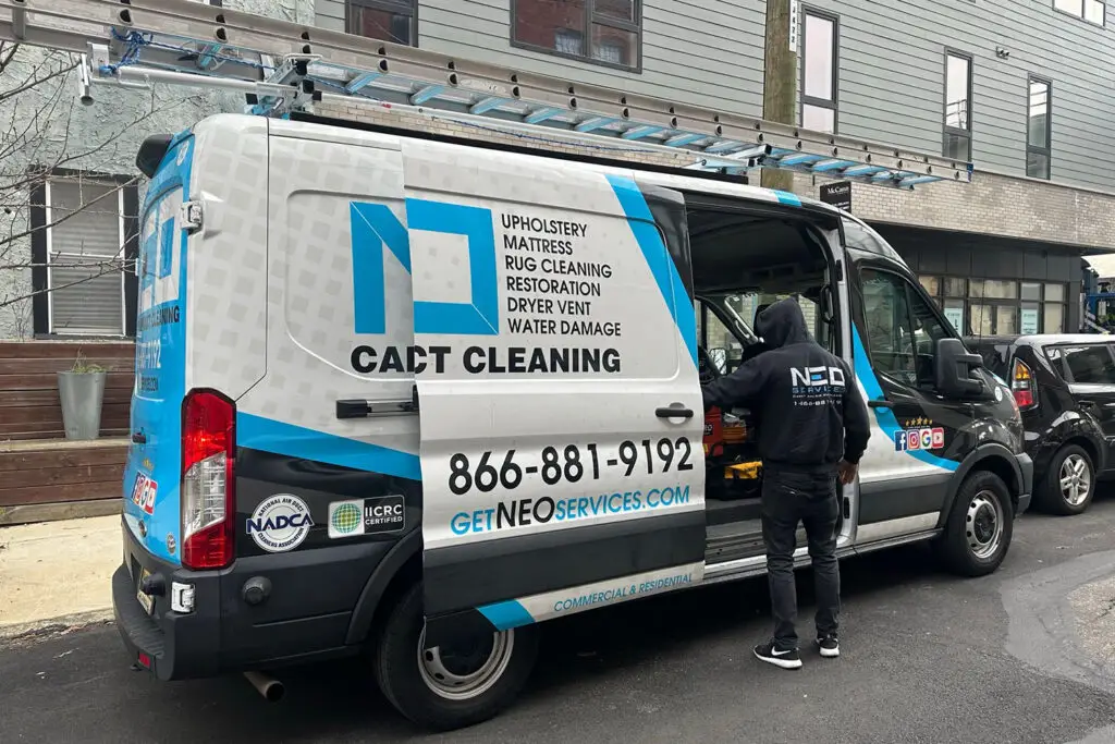 A person in a black hoodie stands next to an open white and blue service van labeled CACT Cleaning. This top-rated van promotes services such as upholstery, mattress, rug, and dryer vent cleaning. It provides contact details including a phone number and website. A ladder is mounted on top.