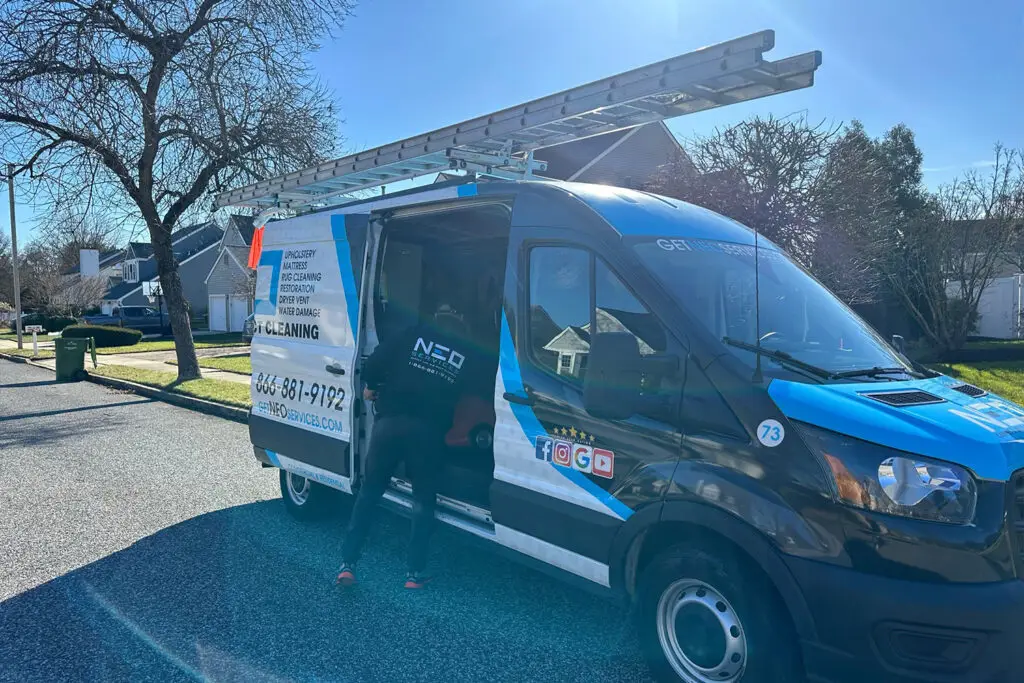 A utility van with a blue and white design, showcasing logos and contact info for a top-rated duct cleaning service, is parked on a residential street. A person steps out as social media icons glint on the side. A ladder rests on top, framed by leafless trees and houses in the background.