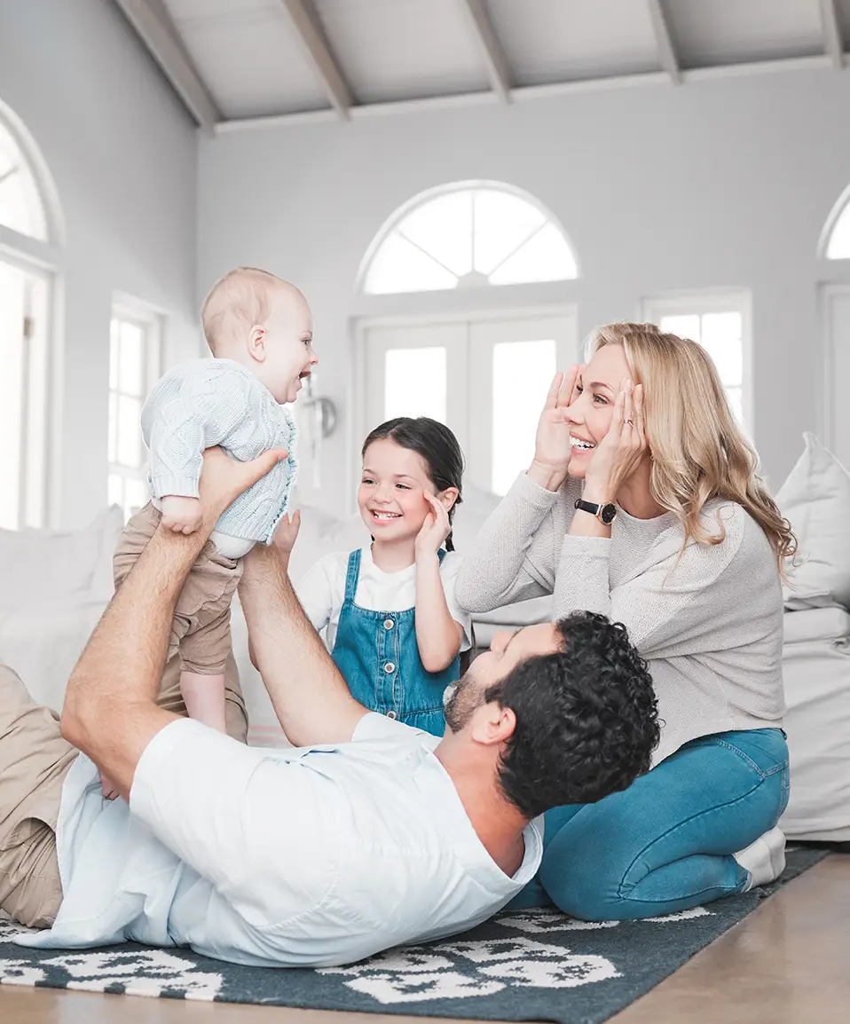 A joyful family moment unfolds in a bright living room: a man lies on the floor holding a smiling baby up in the air. A woman and young girl, sitting nearby, enjoy the playful scene. Thanks to recent air duct cleaning, the homes fresh ambiance enhances the cozy atmosphere of light walls and large windows.