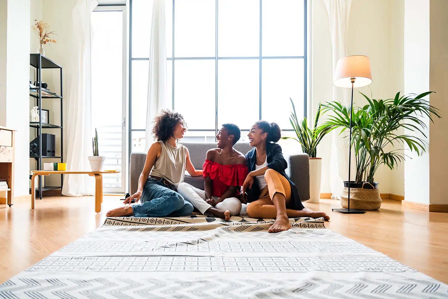 Three people are sitting on a rug in a bright, top-rated living room with large windows. They appear to be laughing and enjoying each others company. The room has plants, a tall lamp, and a shelf with books and decor. The atmosphere is cozy and relaxed, with natural light flooding the space.