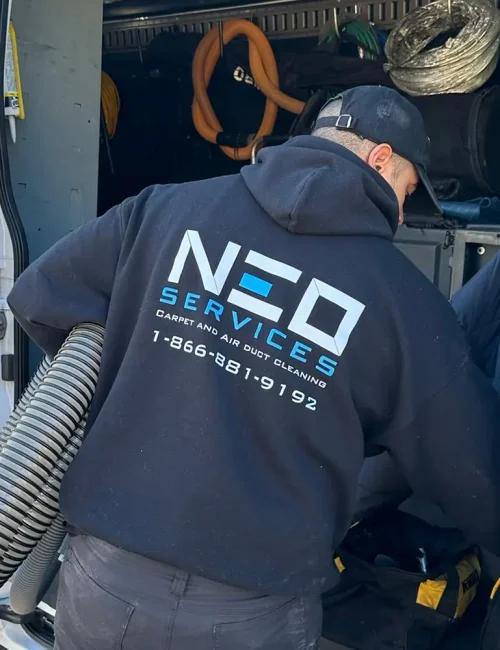 A person wearing a black NEO Services hoodie is working from the back of a van equipped with air duct cleaning tools and large hoses. This setup suggests a restoration service, with the companys contact info and dryer vent cleaning near me featured on the vans side.