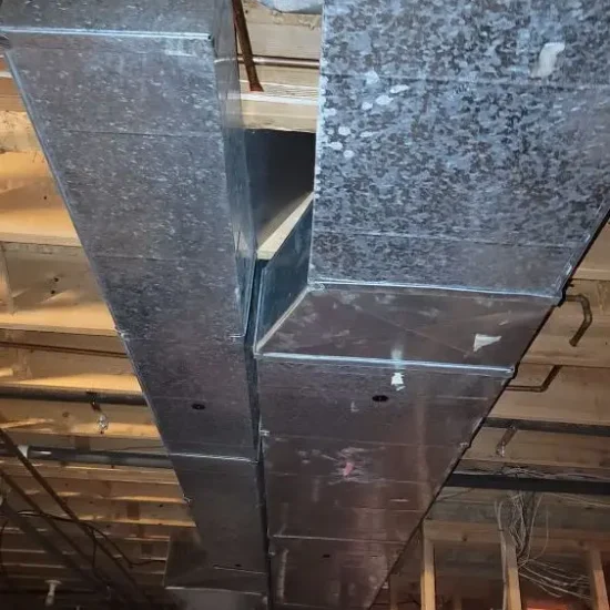 Ceiling view of a basement with exposed wooden beams and aged metal air ducts, hinting at the need for duct cleaning. The ducts run horizontally, showing wear and discoloration. Various pipes and cables are visible against unfinished walls and ceiling insulation, suggesting ongoing construction or renovation.